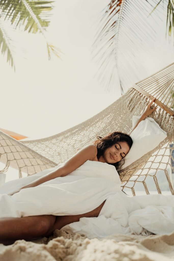 A person relaxing in a hammock on a sandy beach, covered with a white blanket, with palm leaves casting shadows overhead, evoking a tropical, serene vacation vibe.