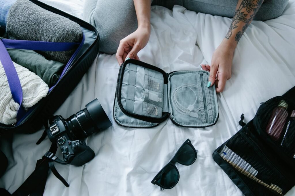 Traveler organizing tech and accessories in a gray packing cube on a white bed, with camera, sunglasses, and suitcase nearby — perfect for carry-on travel in 2025