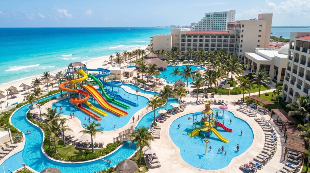 Aerial view of a Cancun beachfront family resort featuring a large water park with colorful slides in orange, yellow, green, and blue, winding swimming pools, children's water play area, white sand beach, turquoise Caribbean Ocean, palm trees, thatched palapas, and multi-story hotel buildings under clear blue sky