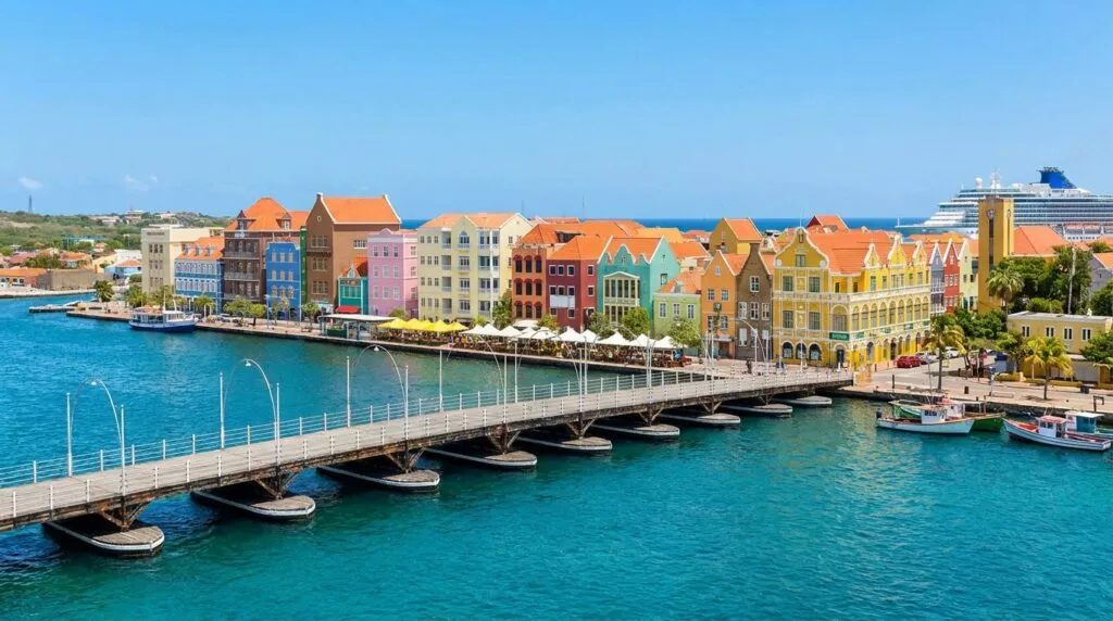 Colorful Dutch colonial buildings in vibrant pink, yellow, orange, blue, and green lining the waterfront of Willemstad, Curaçao, with the iconic Queen Emma floating pontoon bridge crossing turquoise harbor water, cruise ship in background, and palm trees under clear blue sky