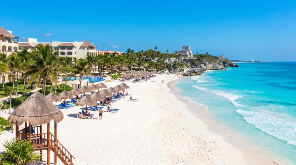 Aerial view of pristine Cancun beach with white sand, turquoise Caribbean water, and thatched palapa umbrellas with blue lounge chairs; luxury beachfront resort with palm trees on the left, and ancient Mayan Tulum ruins perched on rocky limestone cliffs in the background under clear blue sky