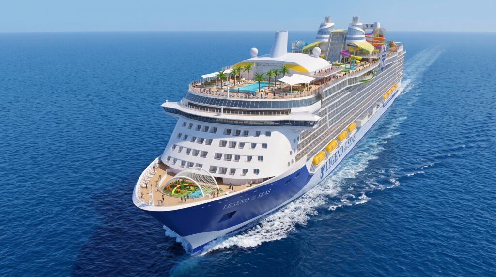 Aerial view of the Royal Caribbean cruise ship Legend of the Seas sailing on deep blue ocean waters, featuring a blue and white hull with multiple passenger decks, colorful water slides in yellow, pink and orange on the upper decks, swimming pools with palm trees, yellow lifeboats along the side, a surf simulator at the bow, and white wake trailing behind the massive vessel under a clear blue sky