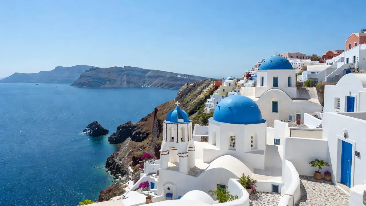 White washed villas with iconic blue domes overlook the deep blue Aegean Sea in Santorini on a sunny day.