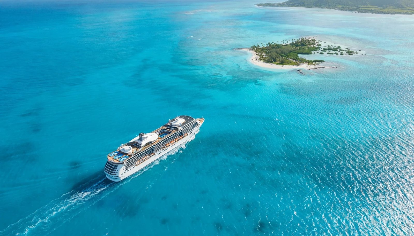 An aerial view of a cruise ship sailing on vivid turquoise water near a small tropical island.