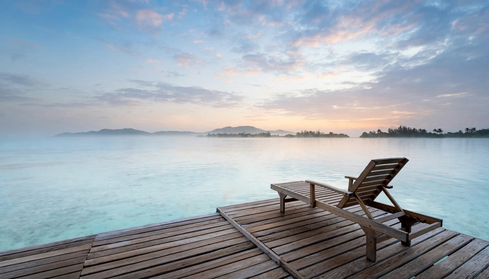An empty wooden lounge chair sits on an overwater deck above a pale Caribbean lagoon, with a distant island under soft dawn light.