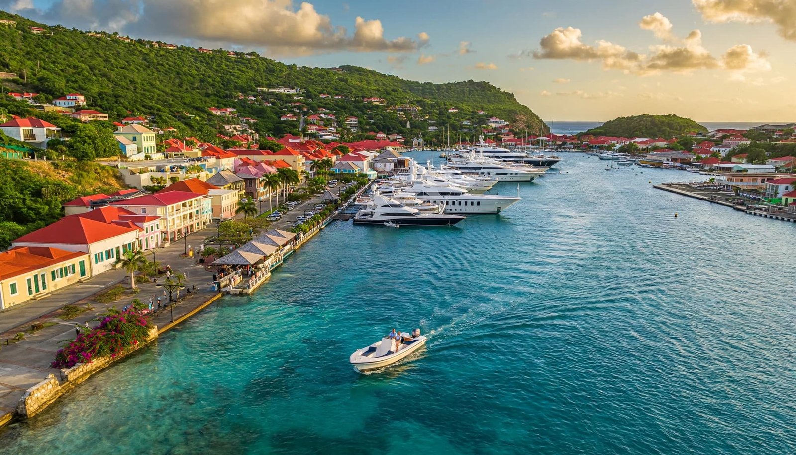 A tender boat passes anchored yachts in Gustavia harbor, with pastel buildings and green hills behind.